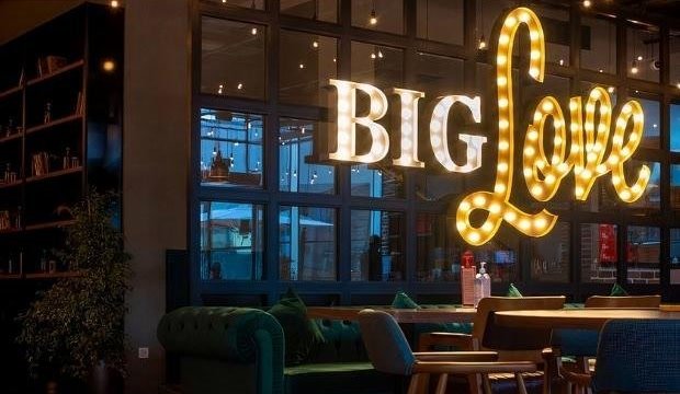 Illuminated “Big Love” marquee letters above a dining room with a green Chesterfield sofa and wooden chairs at BigChefs